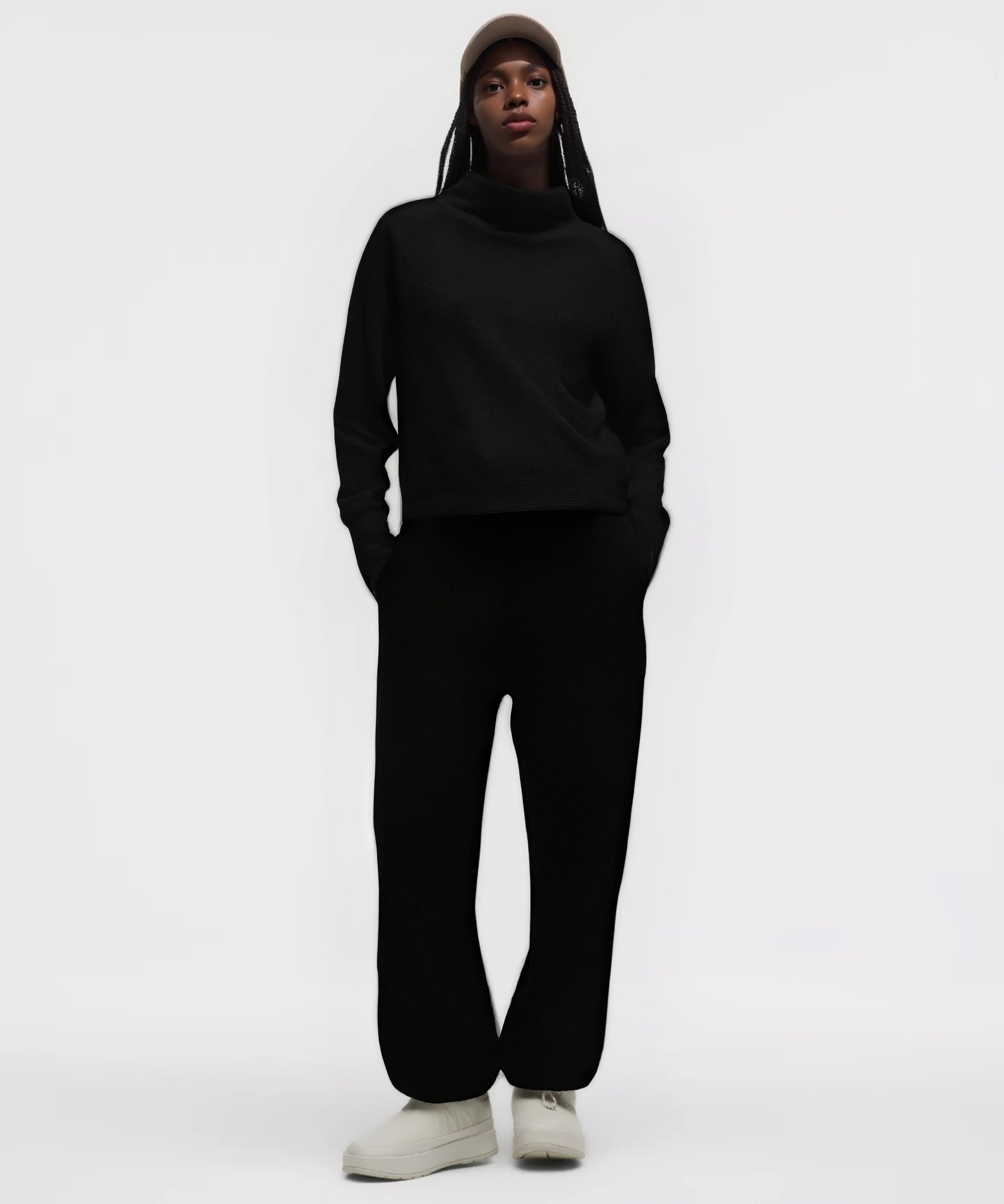 Vue de face d’un mannequin portant l’ensemble noir de la collection GALAYA « SPICA & DENEB », sweat cropped à col montant en texture waffle et pantalon taille haute côtelé, mains dans les poches, posture affirmée, bonnet beige foncé, fond blanc studio, silhouette monolithique noire au contraste élégant.