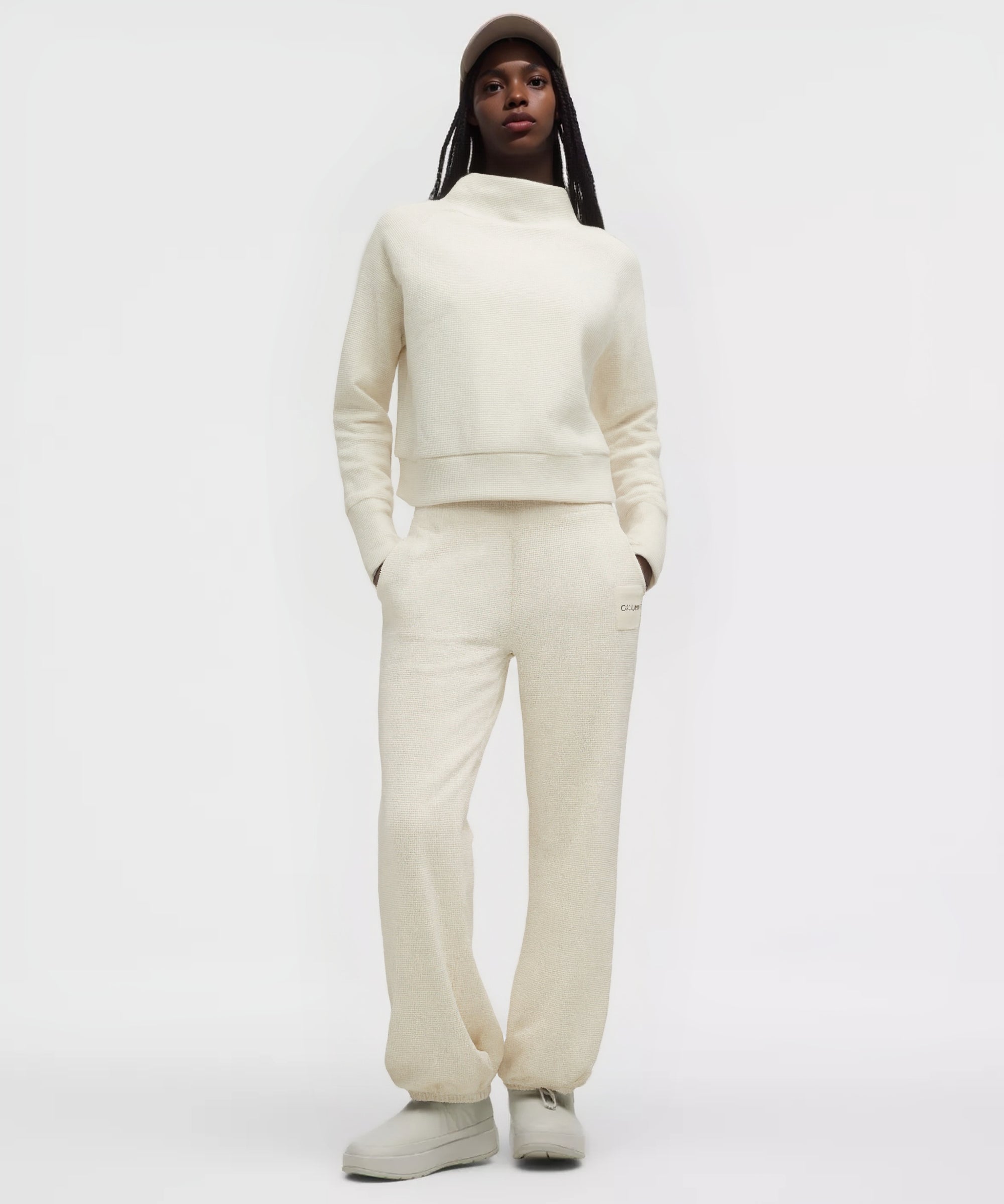 Vue de face d’un mannequin portant l’ensemble ivoire de la collection GALAYA « SPICA & DENEB », sweat cropped à col montant en texture waffle et pantalon taille haute en maille côtelée, mains dans les poches, bonnet beige foncé, fond blanc studio, silhouette minimaliste et affirmée.