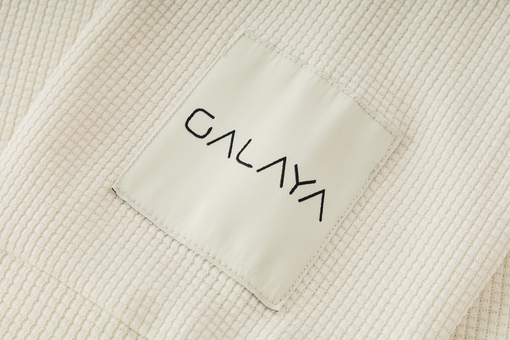 Gros plan sur l’étiquette de la collection GALAYA « SPICA & DENEB », label satin crème cousu sur une maille côtelée ivoire à côtes verticales, logo noir minimaliste, lumière douce soulignant le contraste entre tissu texturé et étiquette lisse.