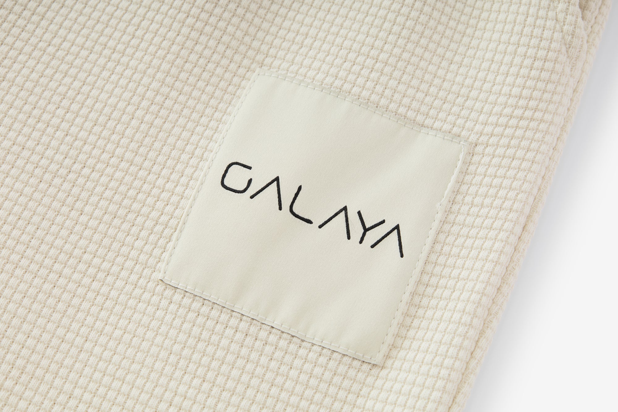 Gros plan sur l’étiquette d’un vêtement de la collection GALAYA « SPICA &amp; DENEB », étiquette crème cousue ton sur ton avec logo noir minimaliste, posée sur un tissu ivoire texturé, lumière douce mettant en valeur la qualité et la discrétion du branding.