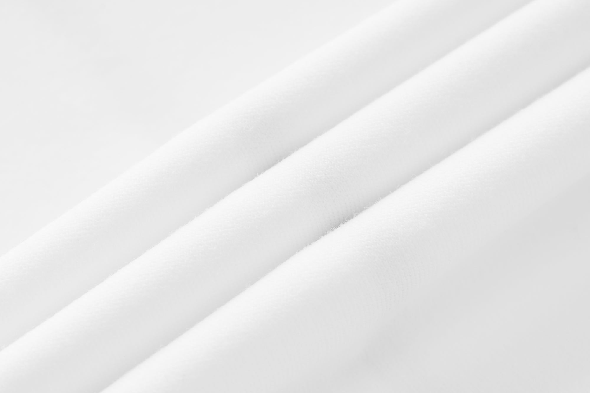 Gros plan macro sur le tissu blanc pur de la collection GALAYA « MIRA &amp; LYRA », jersey lisse et fluide type seconde peau, plis doux et ondulés créant un mouvement naturel, lumière studio très douce et diffuse, surface satinée et uniforme, arrière-plan blanc minimaliste, mettant en valeur la fluidité, la légèreté et la pureté de la matière.